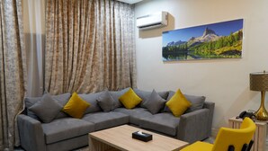 A living room with a grey sectional sofa, yellow cushions, a wooden coffee table, a yellow chair, a lamp, and a painting of a mountain landscape. - Arcade Sinclair (Abuja)