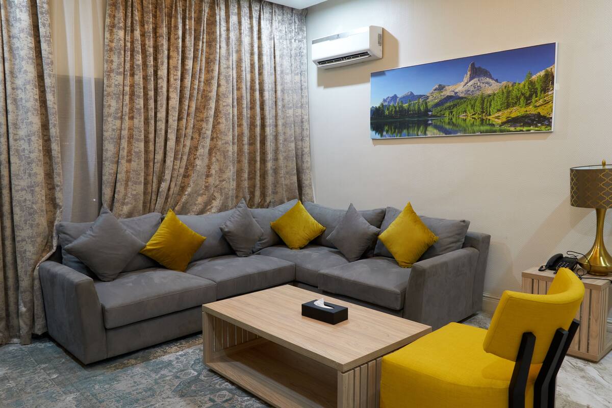 A living room with a grey sectional sofa, yellow cushions, a wooden coffee table, a yellow chair, a lamp, and a painting of a mountain landscape.