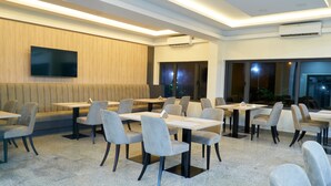 A modern dining area with multiple tables and chairs, a flat-screen TV mounted on the wall, and a large window providing a view of the outdoors. - Arcade Sinclair (Abuja)