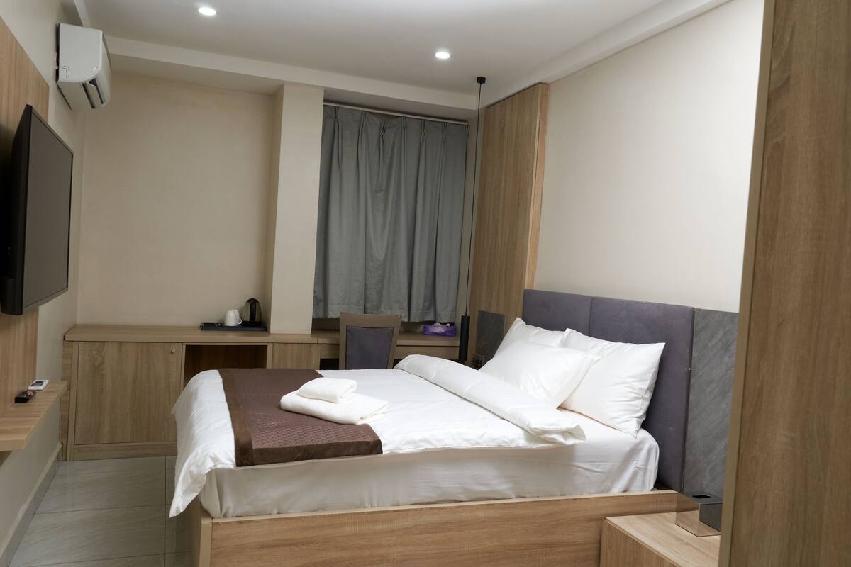 A modern hotel room with a wooden bed, a desk, a chair, a flat-screen TV, and a window with curtains.