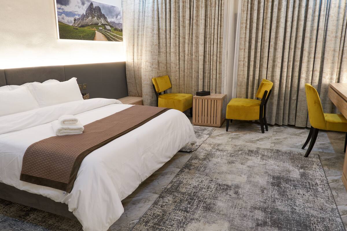 A hotel room with a large bed, a desk, two chairs, a small table, and a view of a mountain through curtains.