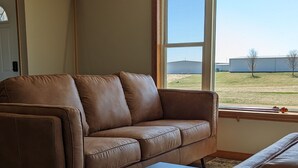 Ein Wohnzimmer mit braunem Ledersofa, einem schwarzen Couchtisch und Blick auf ein grünes Feld durch ein großes Fenster.