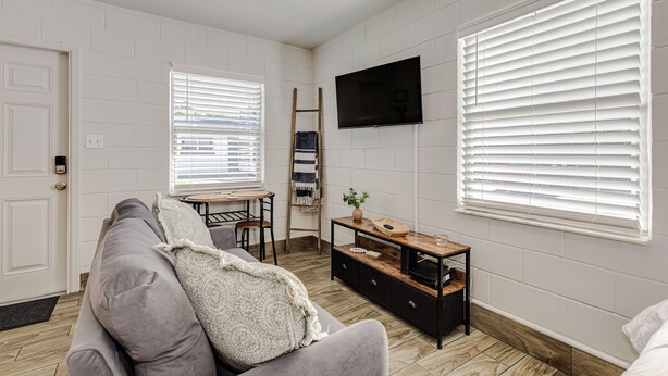A living room with a grey sofa, a wooden coffee table, a TV mounted on the wall, and white blinds on the windows. - Studio Apartment in Winter Haven, FL (Winter Haven)