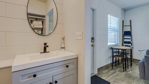 A bathroom with a white vanity, a round mirror, and a towel rack. There is a door leading to a room with a table and chairs. - Studio Apartment in Winter Haven, FL (Winter Haven)