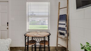 A room with a white brick wall, a window with blinds, a round wooden table with chairs, a wooden ladder, and a TV mounted on the wall. - Studio Apartment in Winter Haven, FL (Winter Haven)