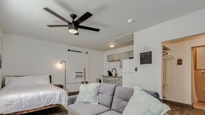 A bedroom with a bed, a gray sofa, a ceiling fan, a nightstand, a wall-mounted lamp, and a small kitchen area with cabinets and a sink. - Studio Apartment in Winter Haven, FL (Winter Haven)