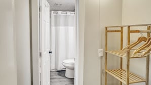 Un couloir étroit mène à une salle de bain équipée de toilettes et d’un rideau de douche. Un meuble en bois avec des cintres se trouve sur la droite.