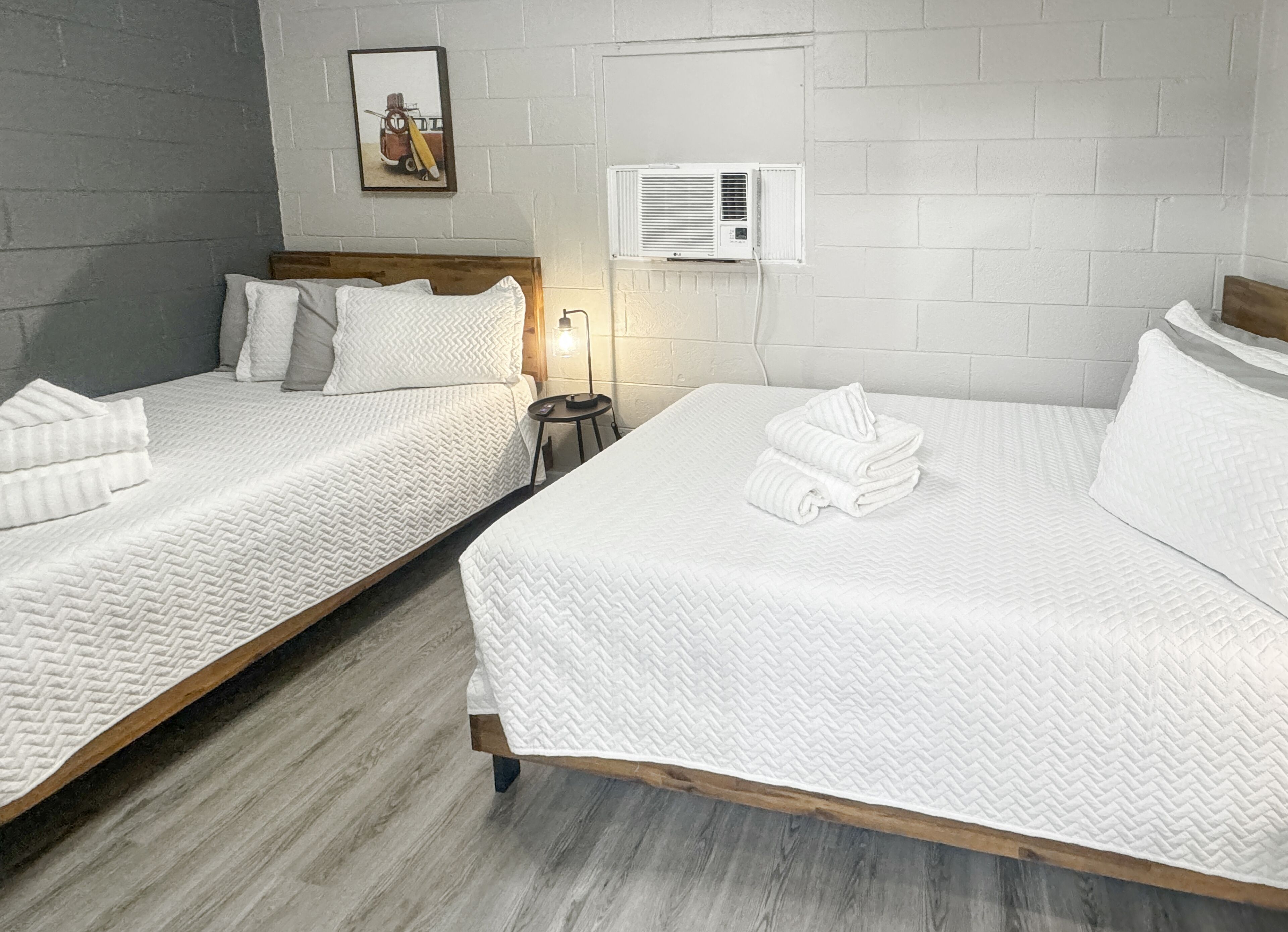 Habitación de hotel con dos camas, cada una con ropa blanca y cabeceros de madera. Hay una mesita con una lámpara y un equipo de aire acondicionado en la pared.