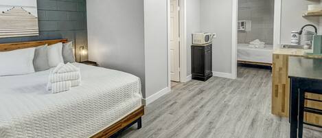 A modern bedroom with a wooden bed, a small kitchenette, and a bathroom visible through an open door.