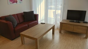A living room with a red sofa, a wooden coffee table, a TV on a wooden stand, and a large window with sheer curtains. - Nest - Lauriedstrasse 9 (Zug)