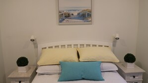 Une chambre à coucher avec un lit blanc, deux tables de chevet avec des lampes, une œuvre d’art encadrée au-dessus du lit et deux petites plantes en pot sur les tables de chevet.