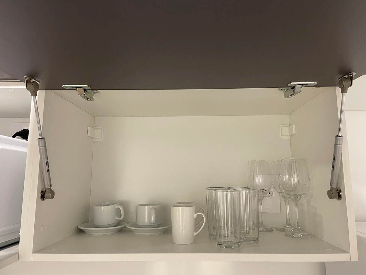 A white cabinet with a shelf containing white cups, saucers, a white mug, and clear wine glasses.