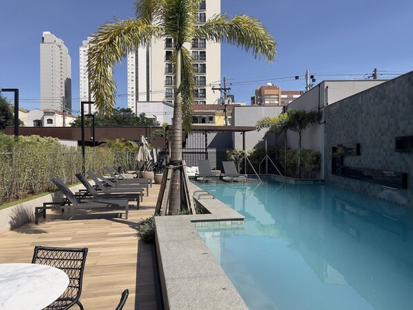 A rooftop pool area with lounge chairs, a palm tree, and a view of tall buildings in the background. - Cyrela For You Carlos Petit (São Paulo)