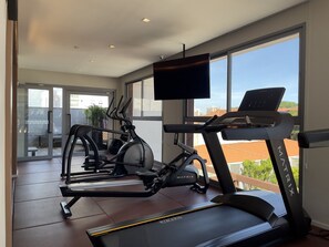 A modern home gym with a treadmill, elliptical, and stationary bike, large windows with a city view, and a flat-screen TV mounted on the wall. - Cyrela For You Carlos Petit (São Paulo)