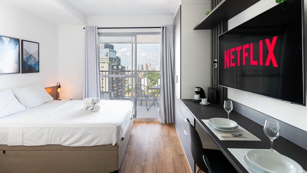 Ein modernes Hotelzimmer mit Bett, Essbereich mit Tellern und Weingläsern, großem Fenster mit Blick auf die Stadt und einem Fernsehbildschirm, der das Netflix-Logo zeigt.