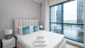 A modern hotel room with a large bed, a nightstand, a lamp, and a view of a cityscape through a large window. - Nasma Holiday Homes - Jumeirah Bay X1 To (Dubai)