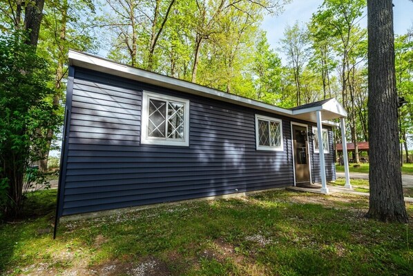 A small, single-story cabin with a dark exterior, two windows with white frames, a porch with a door, and surrounded by trees.