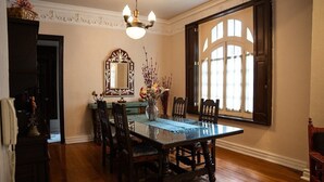 Un comedor con mesa de madera, sillas, una araña, un espejo grande y un ventanal con cortinas.