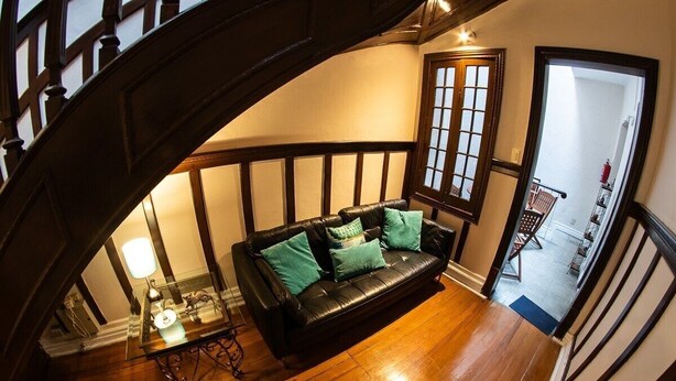 Una escalera conduce a un salón con un sofá de cuero negro y cojines color turquesa, suelo de madera y una mesa de cristal con una lámpara.