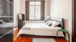 Un dormitorio con cama, una mesita, televisión y ventana con contraventanas.