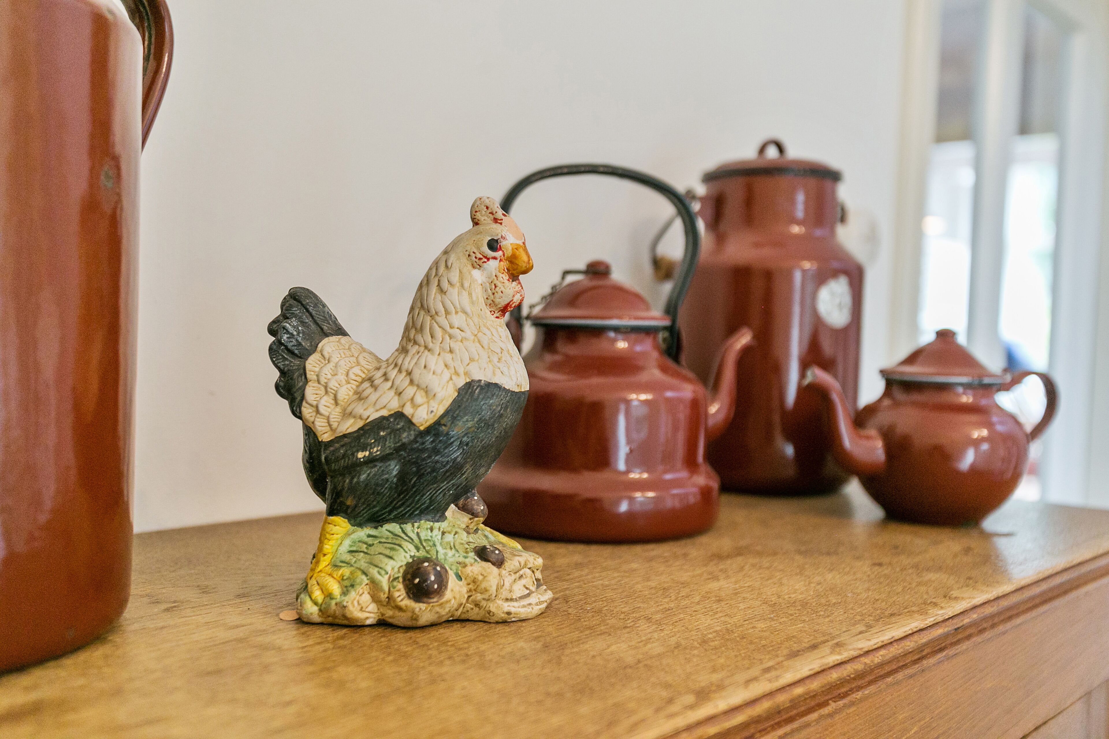A ceramic rooster figurine on a wooden shelf with red pottery items in the background.