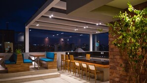 An outdoor seating area with a long table and chairs, a small kitchen area, and a view of the city at night. - Life Residence (Belo Horizonte)