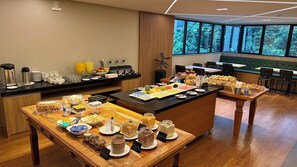 Un bufé de desayuno con diversos alimentos y bebidas, una estación de café y un área de estar con vistas a árboles a través de amplios ventanales.