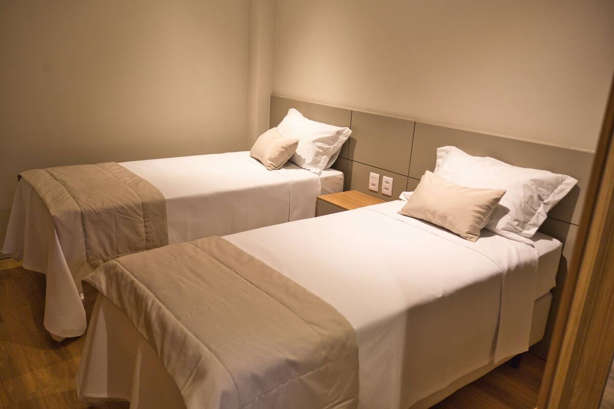Two twin beds with white bedding and beige accents, a wooden nightstand, and a wall-mounted power outlet.