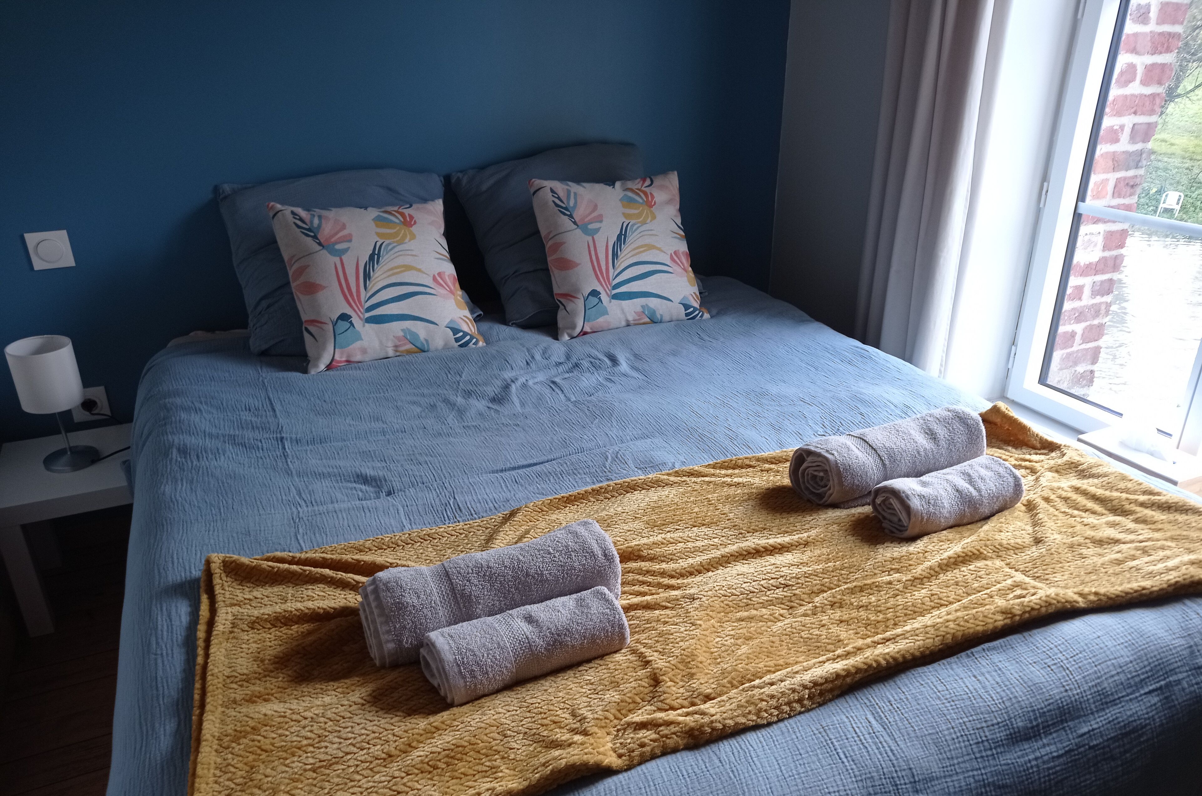 Ein ordentlich bezogenes Bett mit blauer Tagesdecke, zwei Zierkissen, einer gelben Decke und zwei aufgerollten Handtüchern.