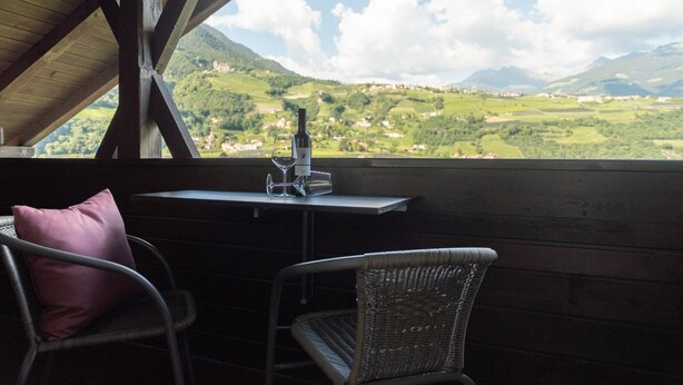 Un balcon avec des fauteuils en osier, une table avec une bouteille de vin et un verre, et une vue sur une vallée verdoyante et des montagnes.