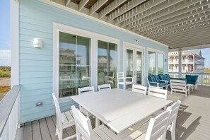 Une terrasse extérieure couverte, avec une table et des chaises blanches, de grandes portes vitrées et une vue sur une maison de plage.