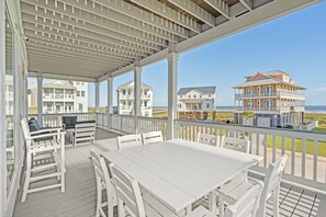Un espace extérieur abrité, avec une table et des chaises blanches, donnant sur une plage où l’on aperçoit des maisons au loin.