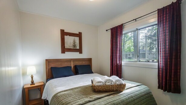 A bedroom with a bed, a nightstand, a lamp, a window with curtains, and a framed picture on the wall. - Cottages #7 Tree Tops, 2 Bedroom Cottage (Lake City)
