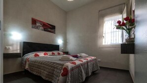 Un dormitorio con cama, mesita de noche, ventana con cortinas y estante en la pared con un jarrón de flores.