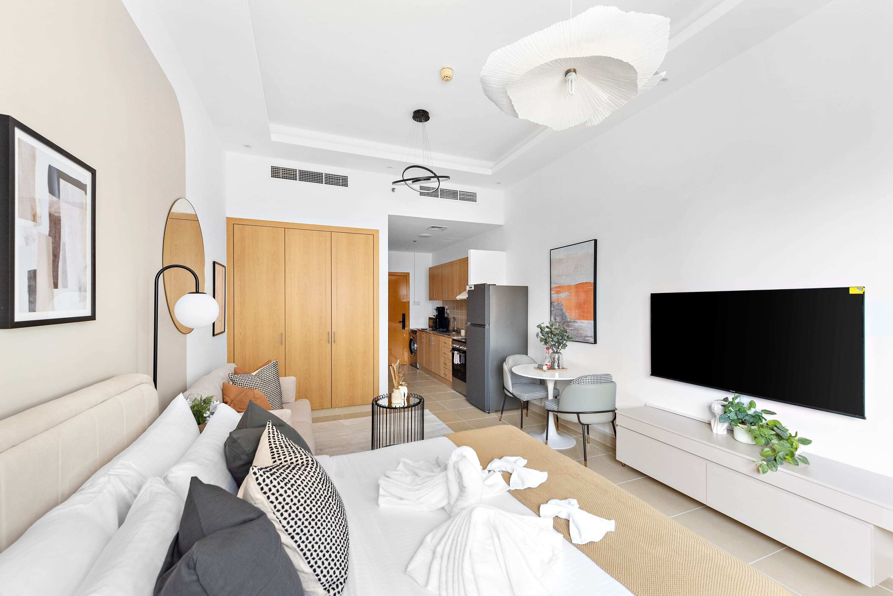 A modern hotel room with a large bed, a TV, a dining area, and a kitchen in the background.