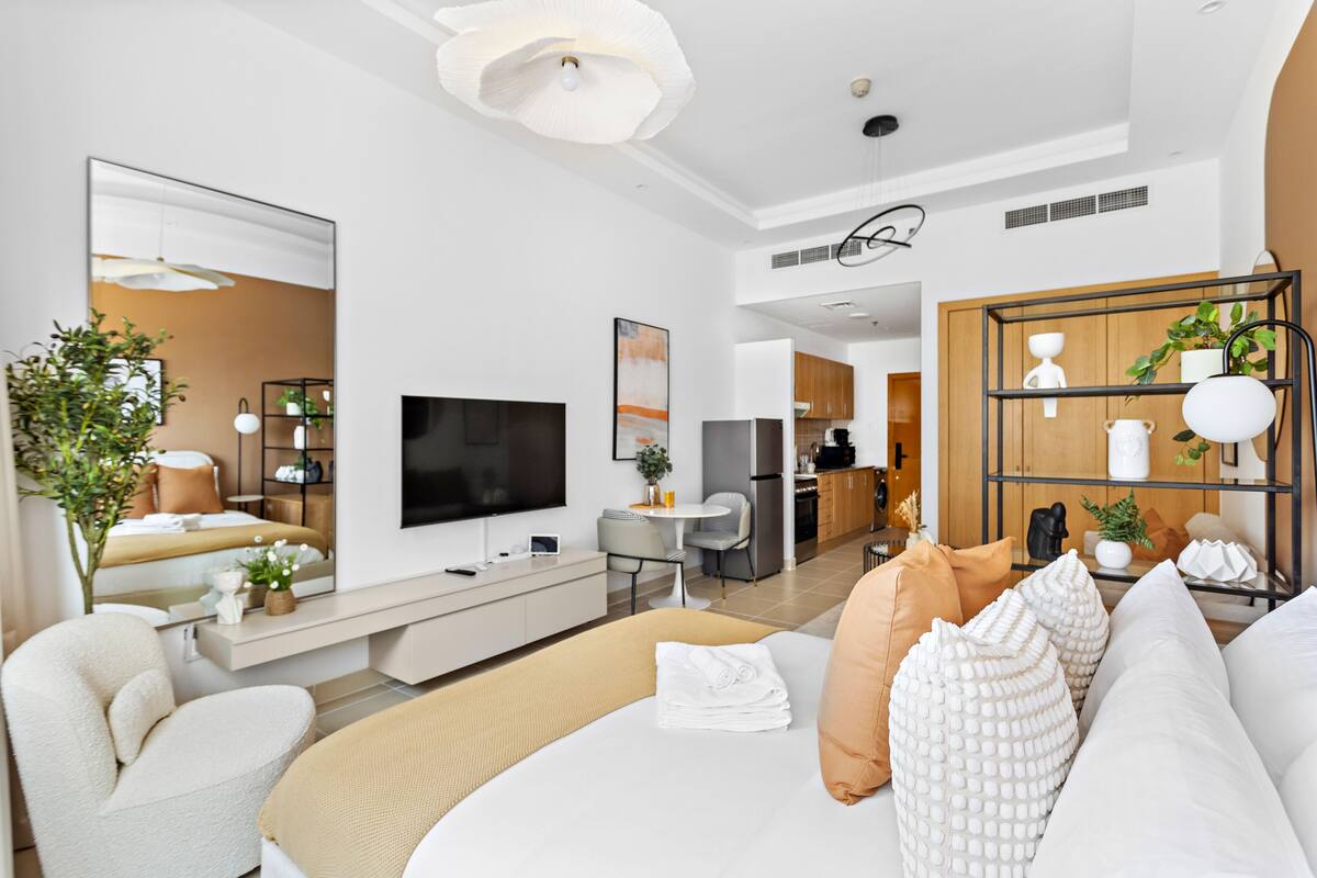 A modern hotel room with a large bed, a flat-screen TV, a seating area with a sofa and armchairs, a dining table, and a kitchen area in the background.