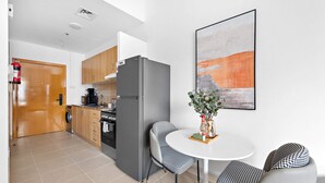 A modern kitchen with a refrigerator, wooden cabinets, and a coffee maker. There is a dining area with a round table and chairs. - Silkhaus ASB Tower (Dubai)