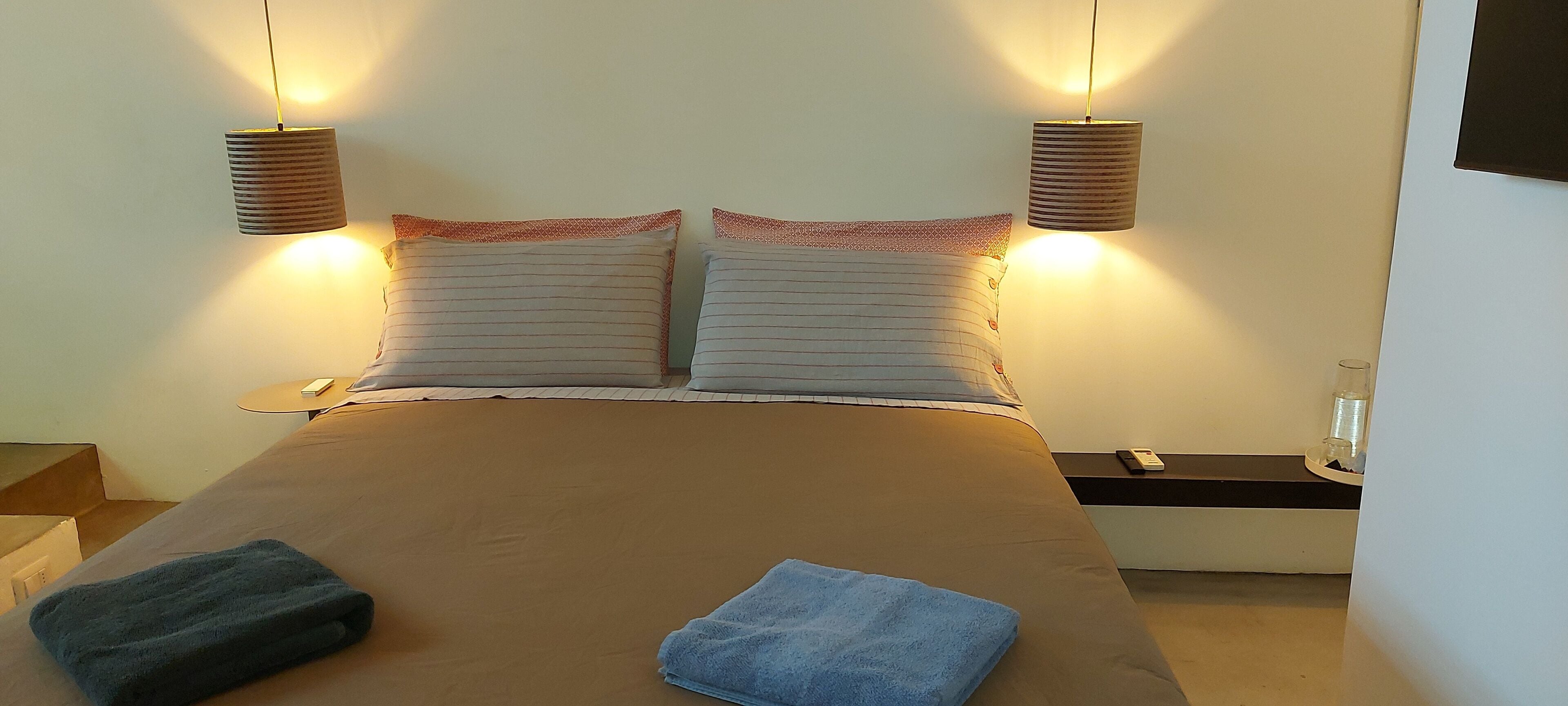 A neatly made bed with two pillows, a blue towel, and a bedside table with a glass and a phone.