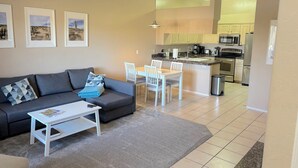 A modern living room with a gray sofa, a white coffee table, and a dining area with white chairs. A kitchen is visible in the background with stainless steel appliances. - McDowell Mountain Adventure Condo - Quiet, Heated Pool, Close to Downtown (Fountain Hills)