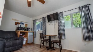 A compact living space with a sofa, a small dining table with chairs, a flat-screen TV mounted on the wall, and a refrigerator.
