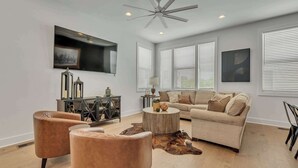 A modern living room with a ceiling fan, a large flat-screen TV, a beige sofa, two leather armchairs, a round wooden coffee table, and a cowhide rug. - 2 Luxury New Homes | Near Broadway + Rooftop Views (Nashville)