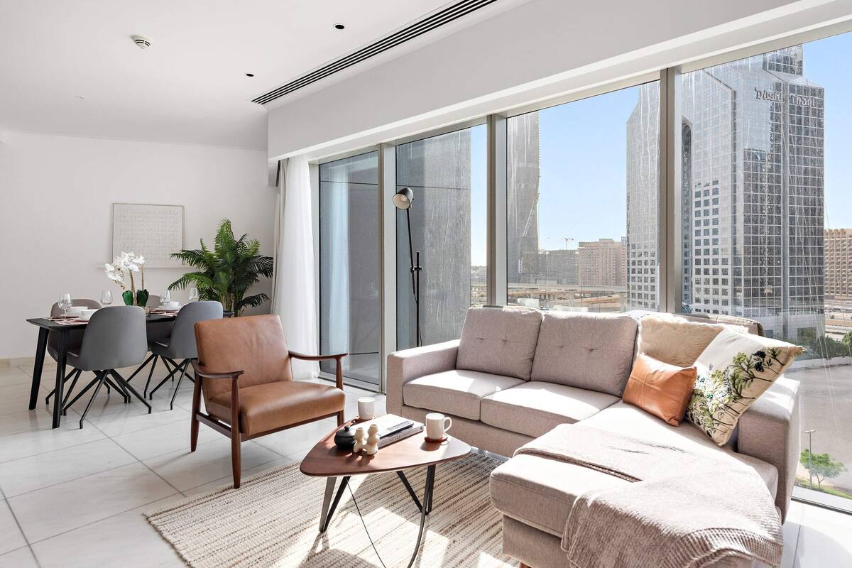 A modern living room with a sofa, armchair, and coffee table, large windows with a city view, and a dining area with chairs and a table.