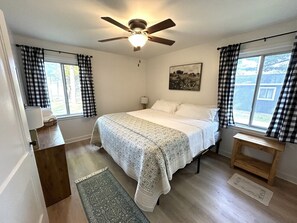 Une chambre à coucher comprenant un lit, un ventilateur de plafond, une fenêtre avec des rideaux, une table de chevet et un tapis.