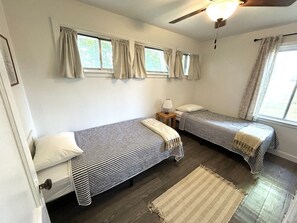 Une chambre avec deux lits simples, un ventilateur de plafond, une fenêtre avec des rideaux, une table de chevet avec une lampe et un tapis sur le sol.