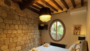 Une chambre avec un mur en pierre, un sol à carreaux, un lit avec des serviettes, une fenêtre ronde et un luminaire suspendu.