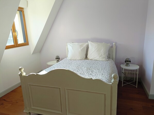 A bedroom with a sloped ceiling, a bed with a floral bedspread, two bedside tables with lamps, and a window with a view of the outside. - Grande Maison, 5 Minutes Centre Honfleur, vue Pont de Normandie (Honfleur)