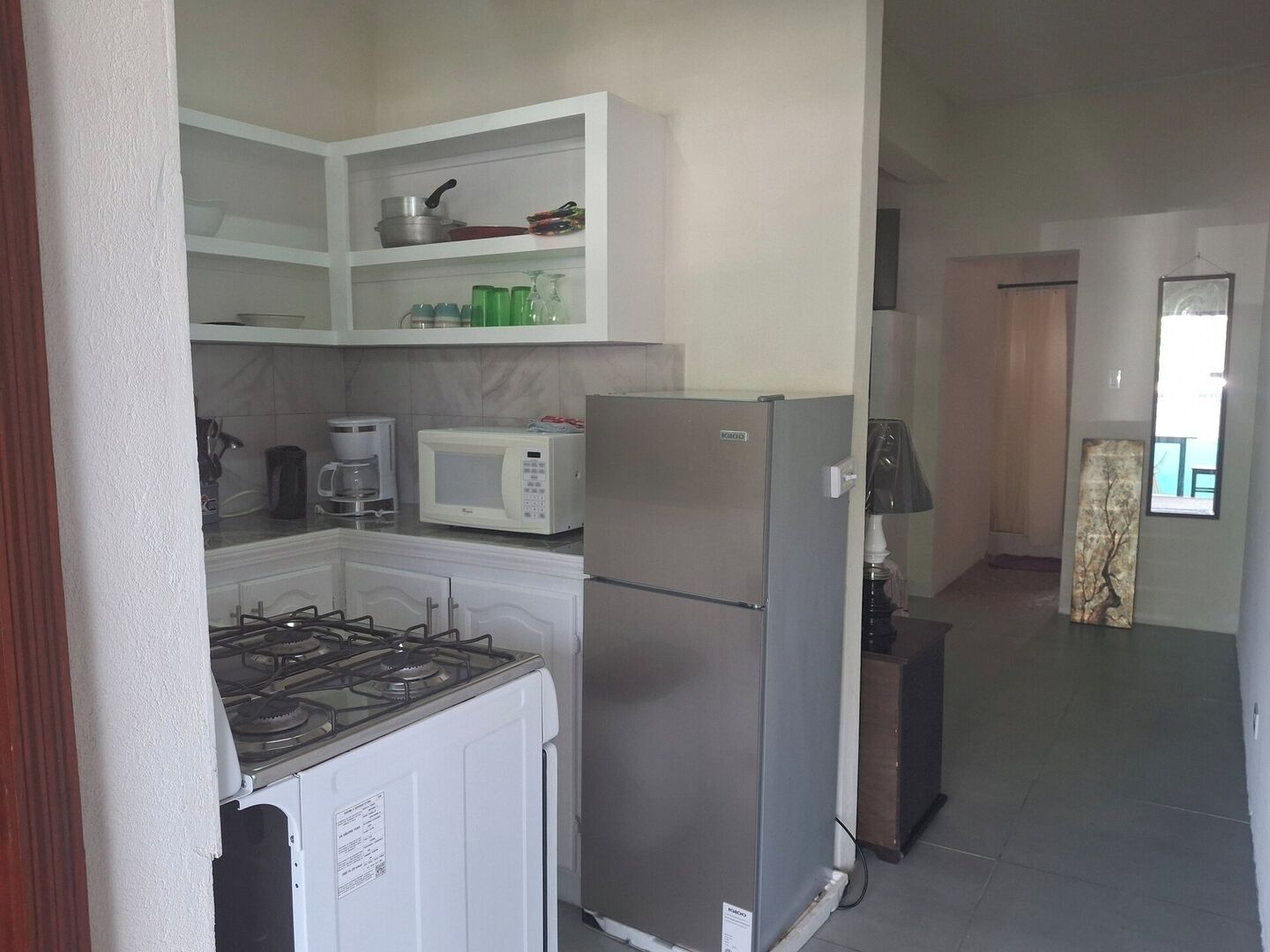A compact kitchen with a gas stove, a microwave, and a refrigerator. There is a shelf above the stove with various kitchen items.