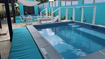 Une piscine dont le bord est revêtu de carreaux, entourée d’une terrasse couverte aménagée avec des sièges et une petite table.