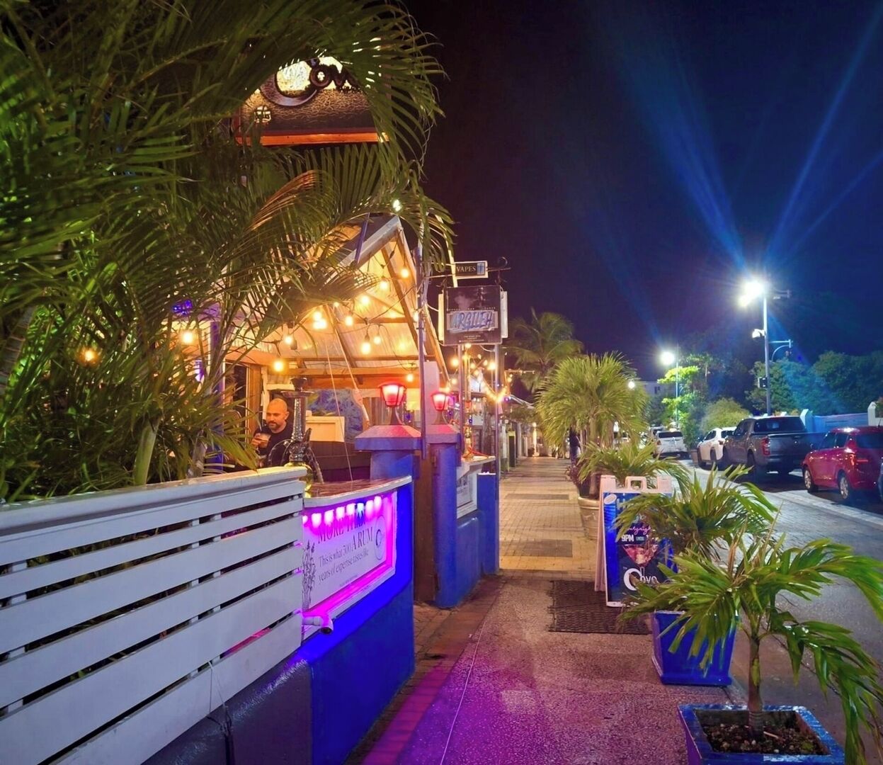 Een restaurant buiten, 's nachts open, met een overdekte zithoek, verlicht door lichtslingers en omgeven door palmbomen.