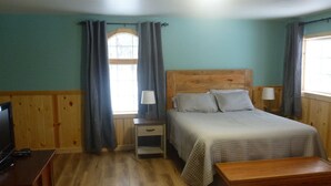 Une chambre à coucher avec une tête de lit en bois, un lit avec des oreillers, une table de chevet avec une lampe, un banc en bois et un téléviseur posé sur une commode en bois.